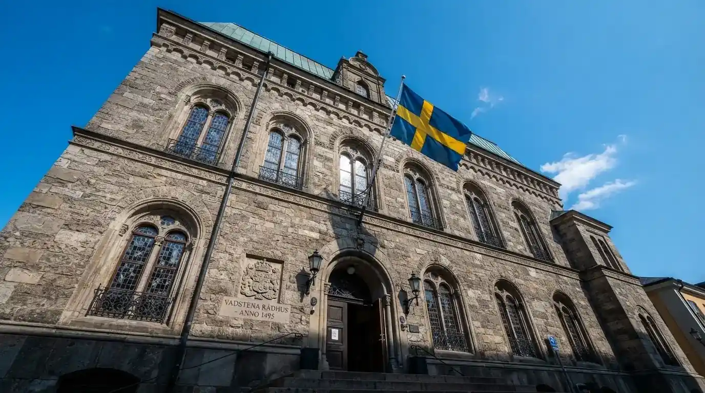 Ett gammalt stadshus i sten fotograferat underifrån mot en klarblå himmel, med den svenska flaggan som fladdrar ovanför entrén.
